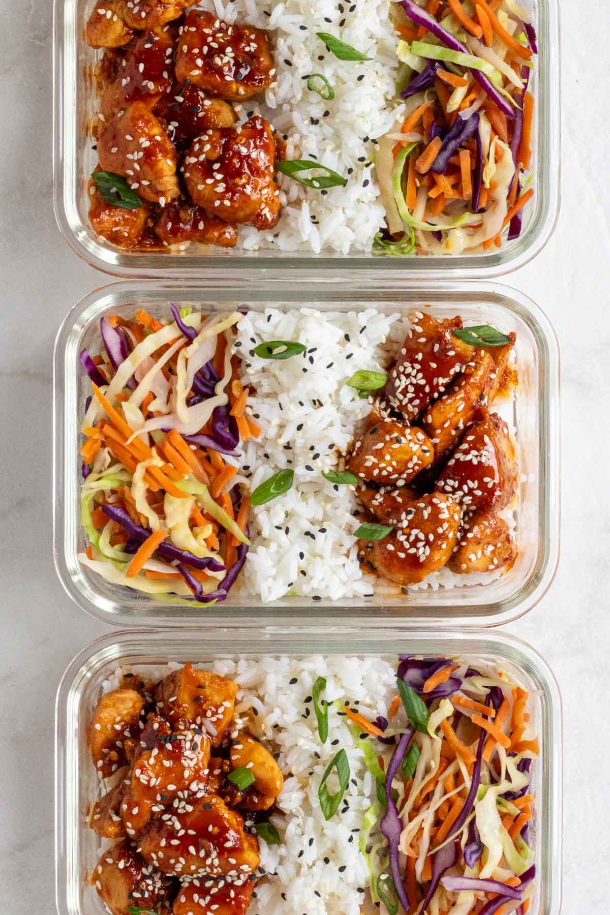 Close of three meal prep sticky chicken bowls with white rice and sautéed shredded cabbage and carrots with sesame seeds and chopped green onions on a white background.
