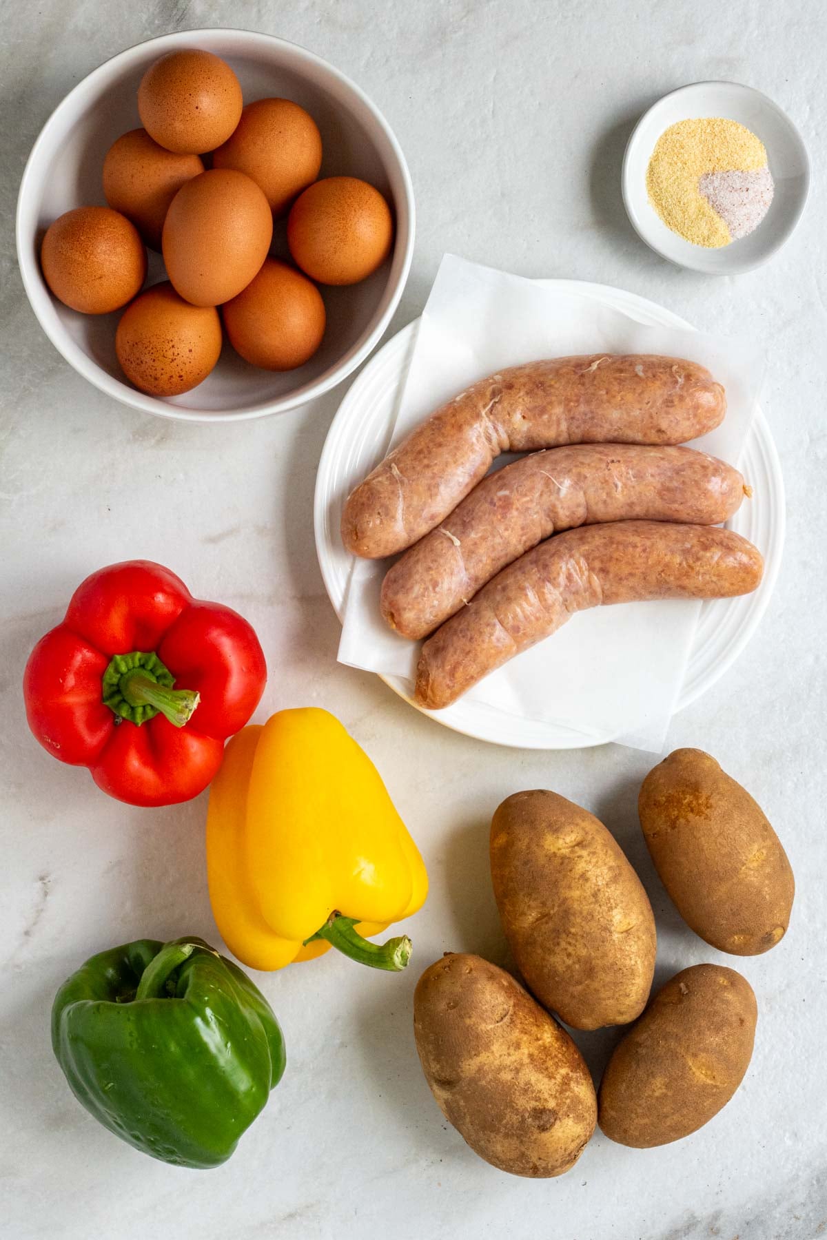 Bowl of eggs, bowl of onion powder, garlic powder, and salt, plate of turkey sausages, bell peppers, and potatoes on a white background.