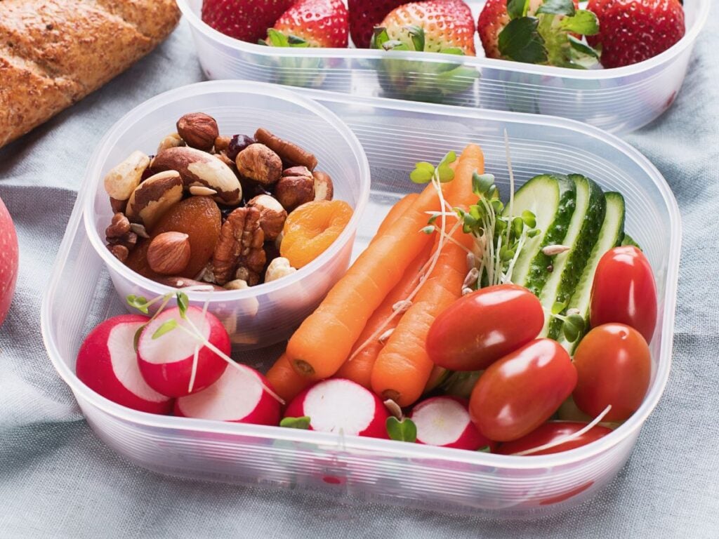 Meal prep container with baby carrots, cucumber, cherry tomatoes, and radishes with a small container of trail mix on a grey tea towel with a container of strawberries in the background and a loaf of bread and apple on the side.