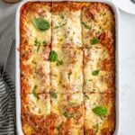 Casserole dish with ricotta and vegetable lasagna sliced into pieces with parmesan cheese, basil, tea towel, spatula, plate and fork on the side.