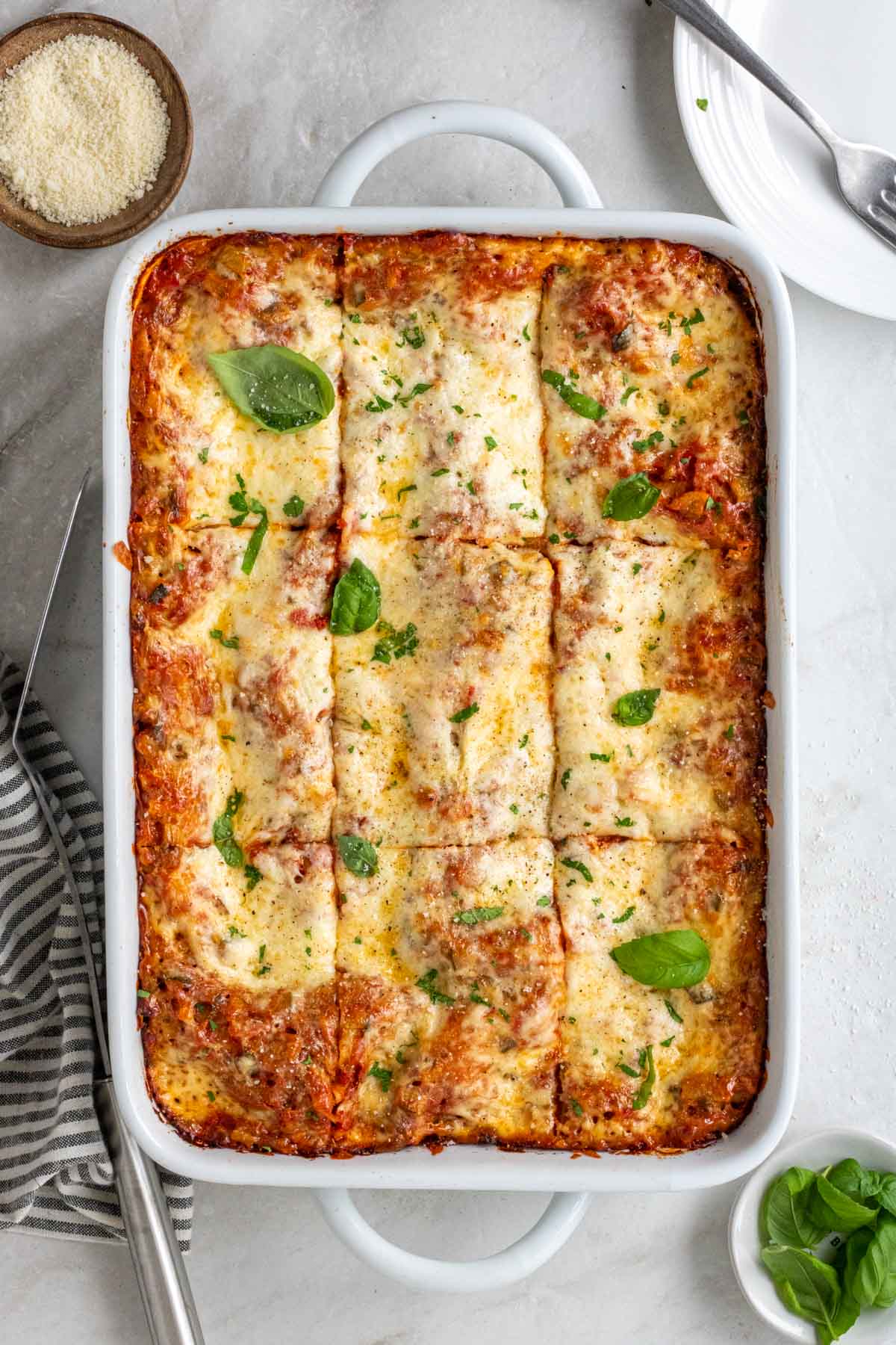 Casserole dish with ricotta and vegetable lasagna sliced into pieces with parmesan cheese, basil, tea towel, spatula, plate and fork on the side.