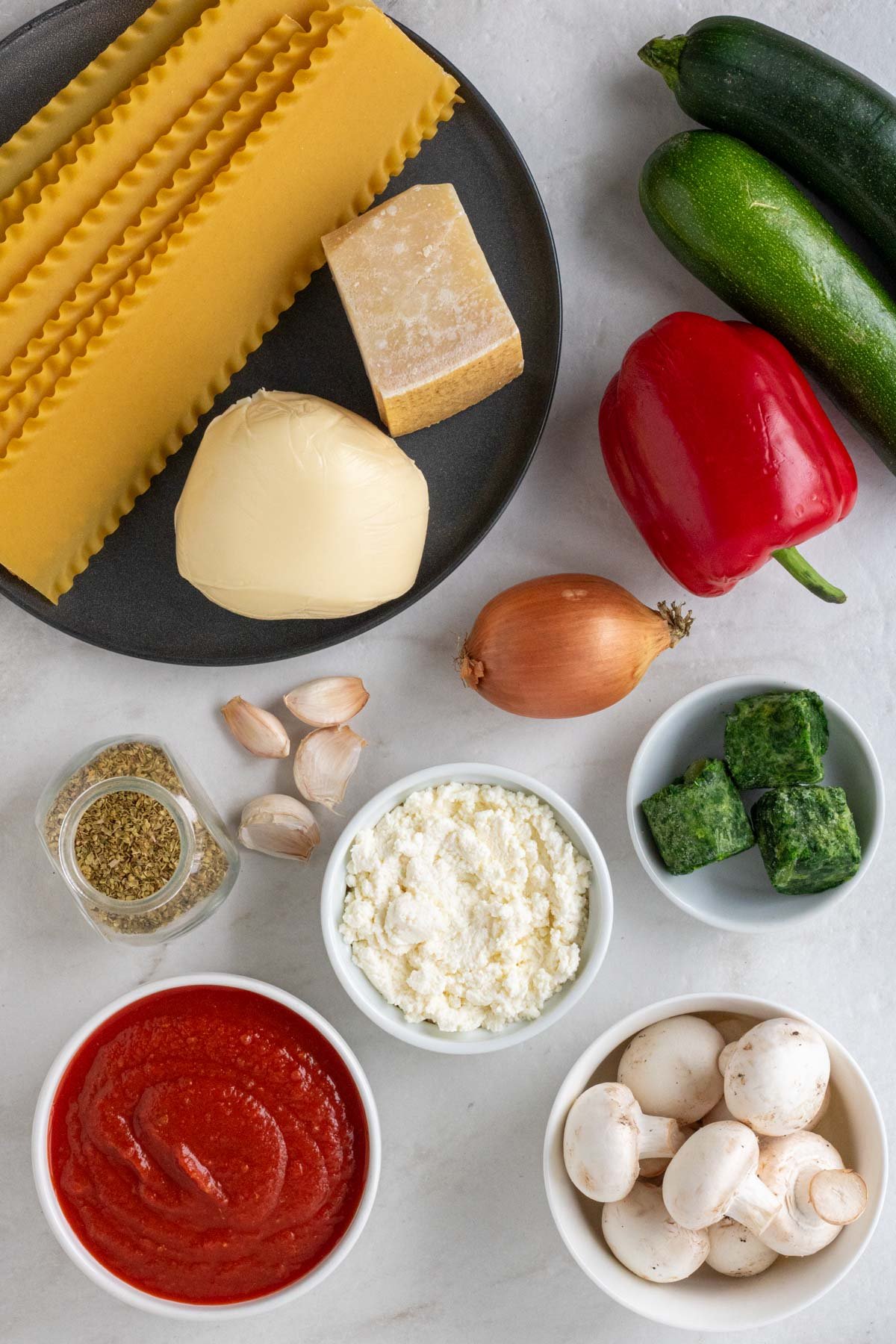 Ingredients with for veggie lasagna: zucchini, red bell pepper, mushrooms, onion, garlic, crushed tomatoes, lasagna noodles, ricotta cheese, frozen spinach, mozzarella cheese, parmesan cheese, egg, oregano, nutmeg, salt, and pepper.
