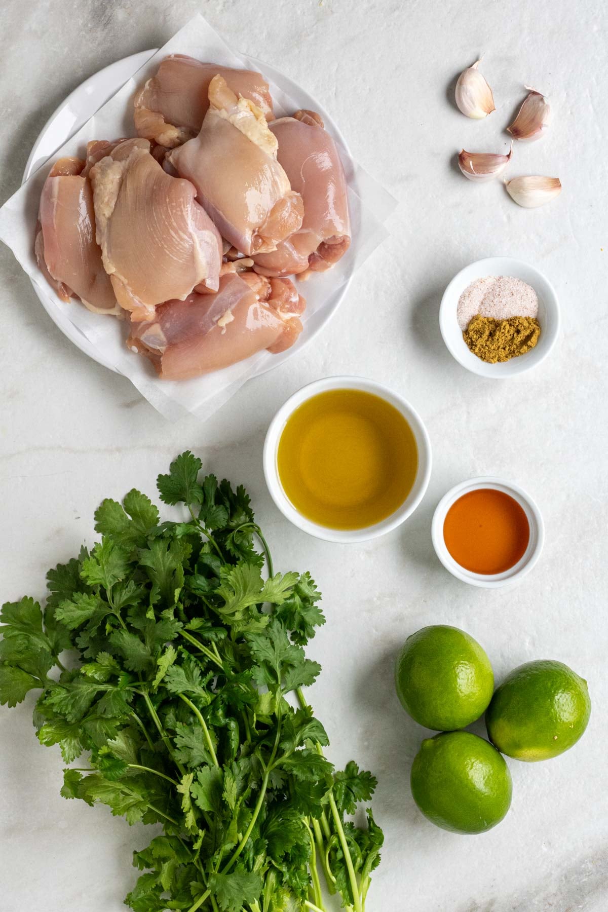Plate of raw chicken thighs, garlic cloves, bowl of salt and cumin, olive oil, honey, fresh cilantro, and limes on a white background.