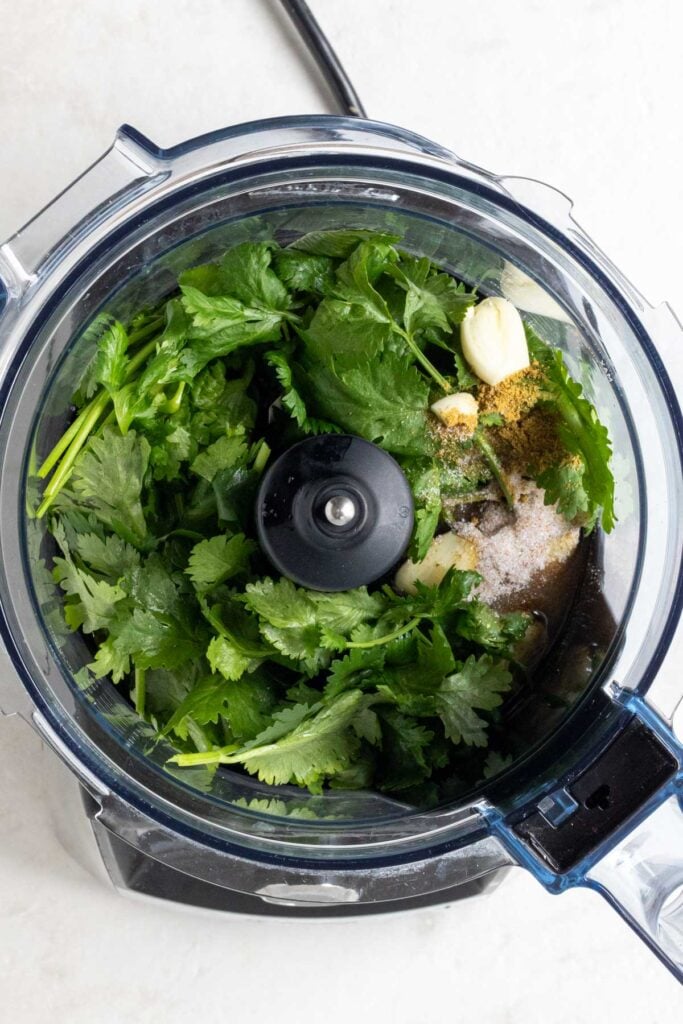 Food processor with olive oil, fresh cilantro, lime juice, garlic cloves, honey, cumin, salt, and pepper.