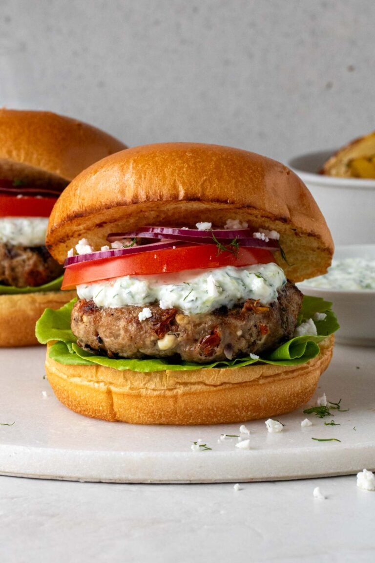 Two Greek chicken burgers with tzatziki, lettuce, sliced tomato, sliced red onion, and crumbled feta cheese on a white cutting board with lemon roasted potatoes in the background.