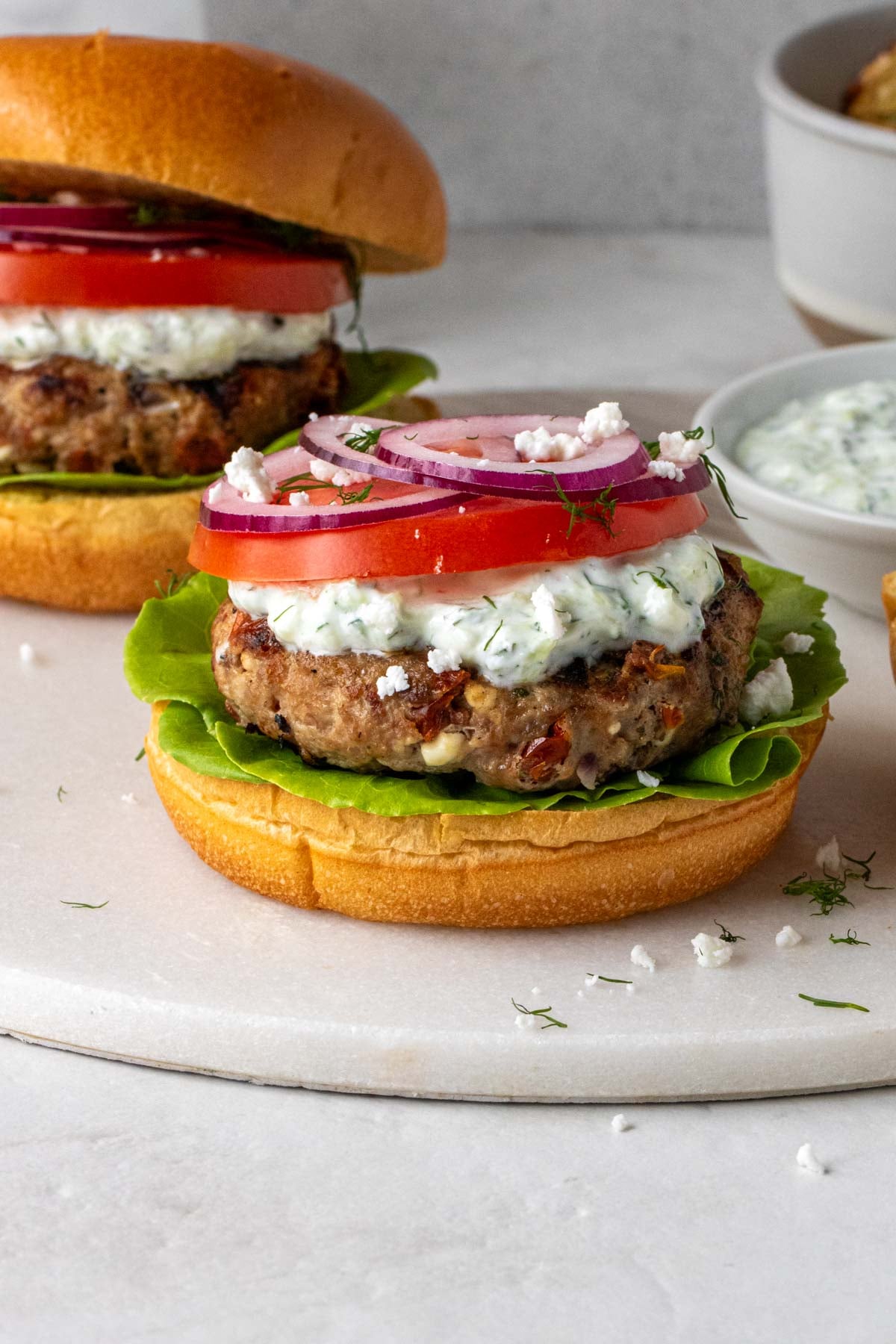 Close up Greek chicken burger on a brioche bun with toppings and a bowl tzatziki in the background on a white cutting board.