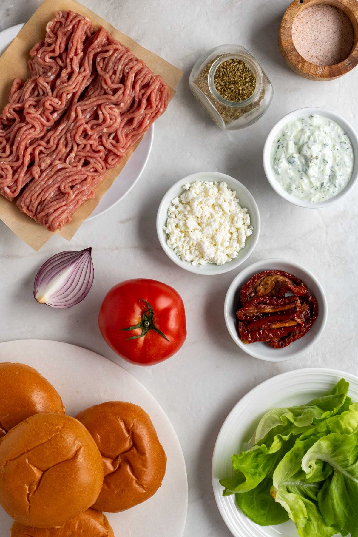 Ground chicken on a white plate with dried oregano, salt, tomato, red onion, bowls of feta cheese, tzatziki sauce, and sun-dried tomatoes on the side with a plate of brioche buns and leafy lettuce on a plate on a white background.