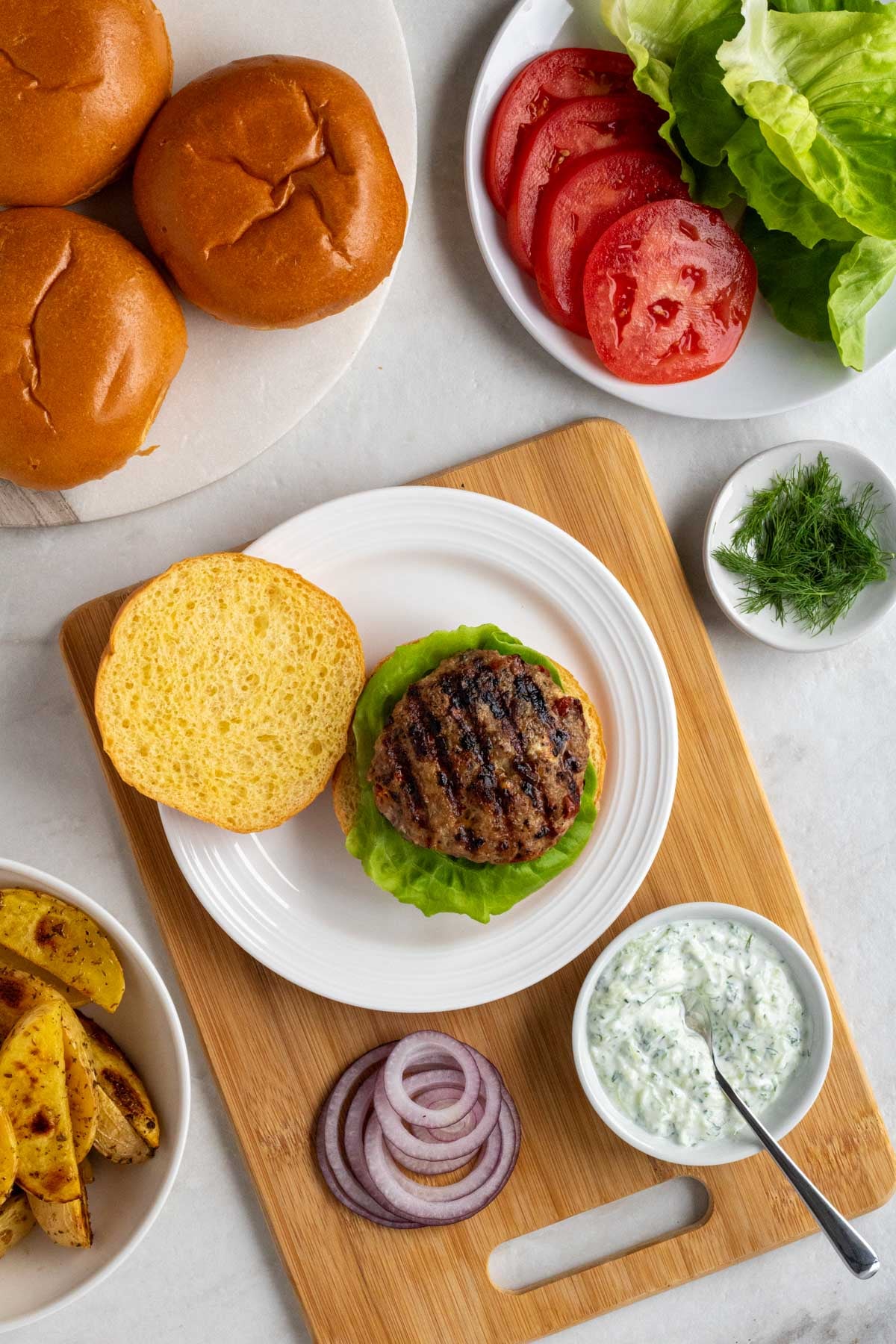 Cooked Greek turkey burger on a plate on a cutting board with a bun with toppings and roasted lemon garlic potatoes on the side.