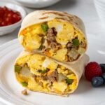 Meal prep breakfast burritos on a white plate with fresh berries on the side and small bowls of salsa and sour cream in the background.