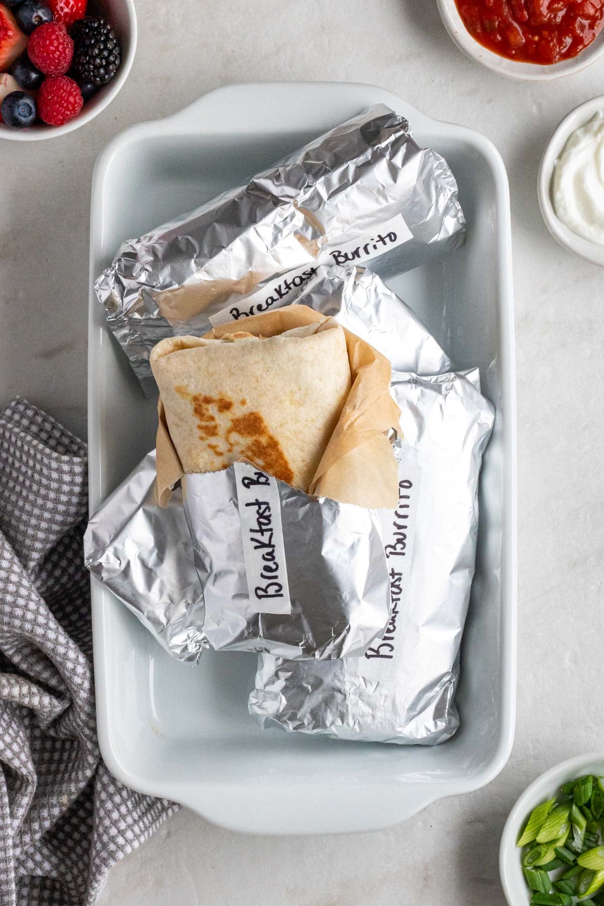 Baking dish of freezer-friendly breakfast burritos wrapped in aluminum foil with a grey tea towel and bowls of fresh berries, salsa, sour cream, and slice green onions on a white background.
