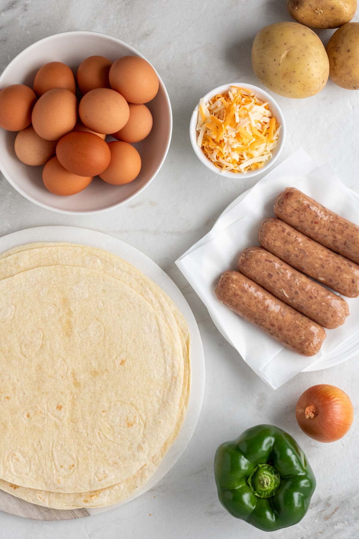 White bowl of eggs, potatoes, bowl of shredded cheese, plate of sausages, plate of tortillas, onion, and green pepper on a white background.