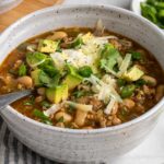 Close up of a bowl of white bean turkey chili with diced avocado, shredded cheese, cilantro, and green onion on top with a silver spoon on a blue and white striped tea towel on a white background.