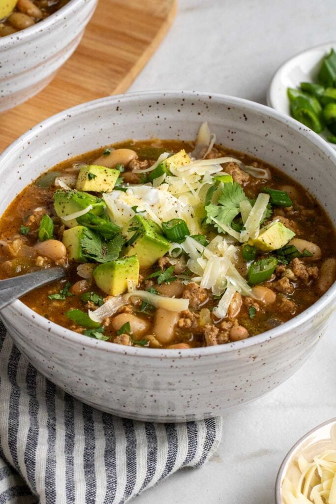 Close up of a bowl of white bean turkey chili with diced avocado, shredded cheese, cilantro, and green onion on top with a silver spoon on a blue and white striped tea towel on a white background.