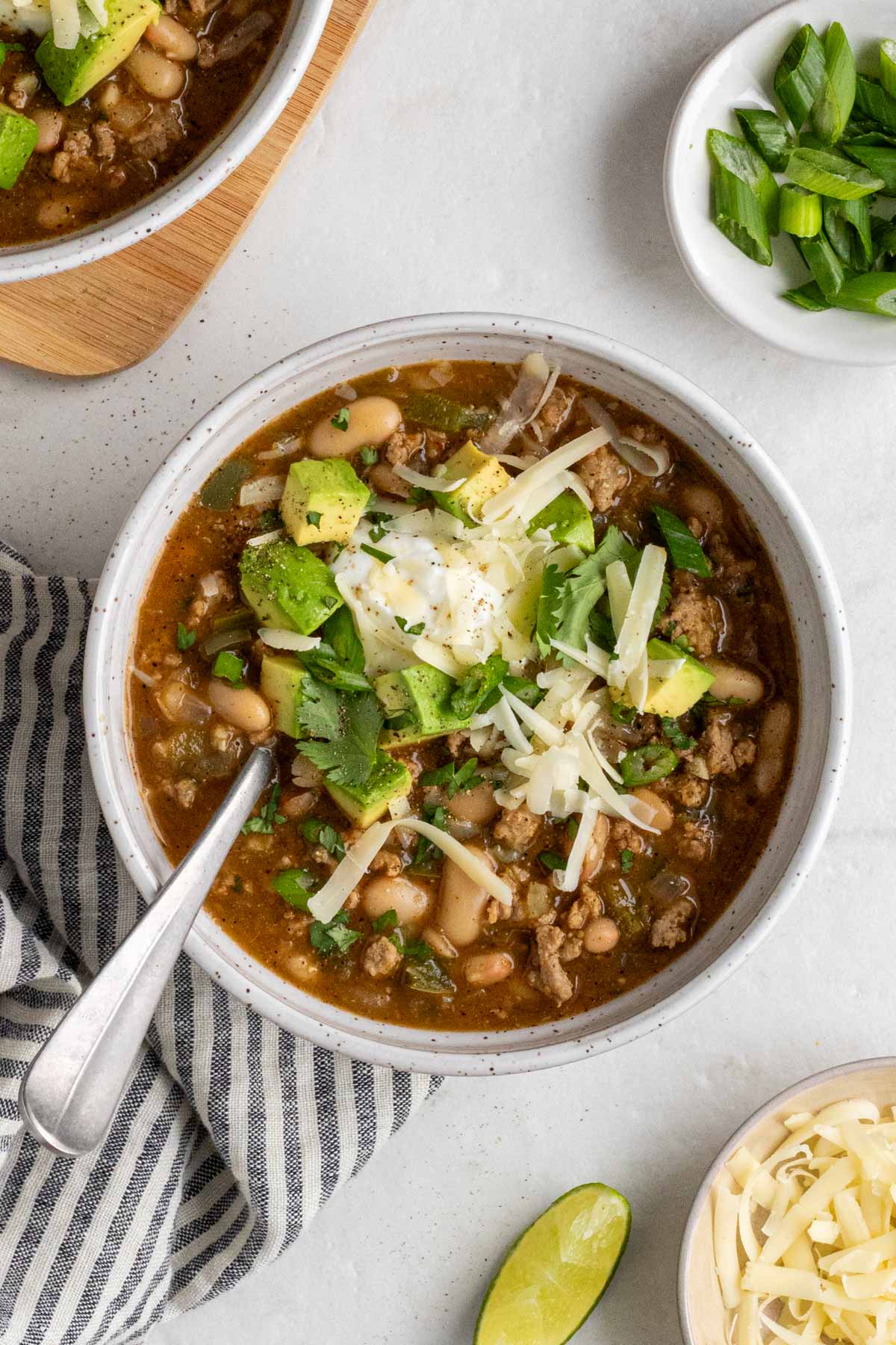 Two white speckled bowls of white bean turkey chili with small bowls of sliced green onion and shredded cheese and a lime wedge on a white background with a stripped blue and white tea towel.