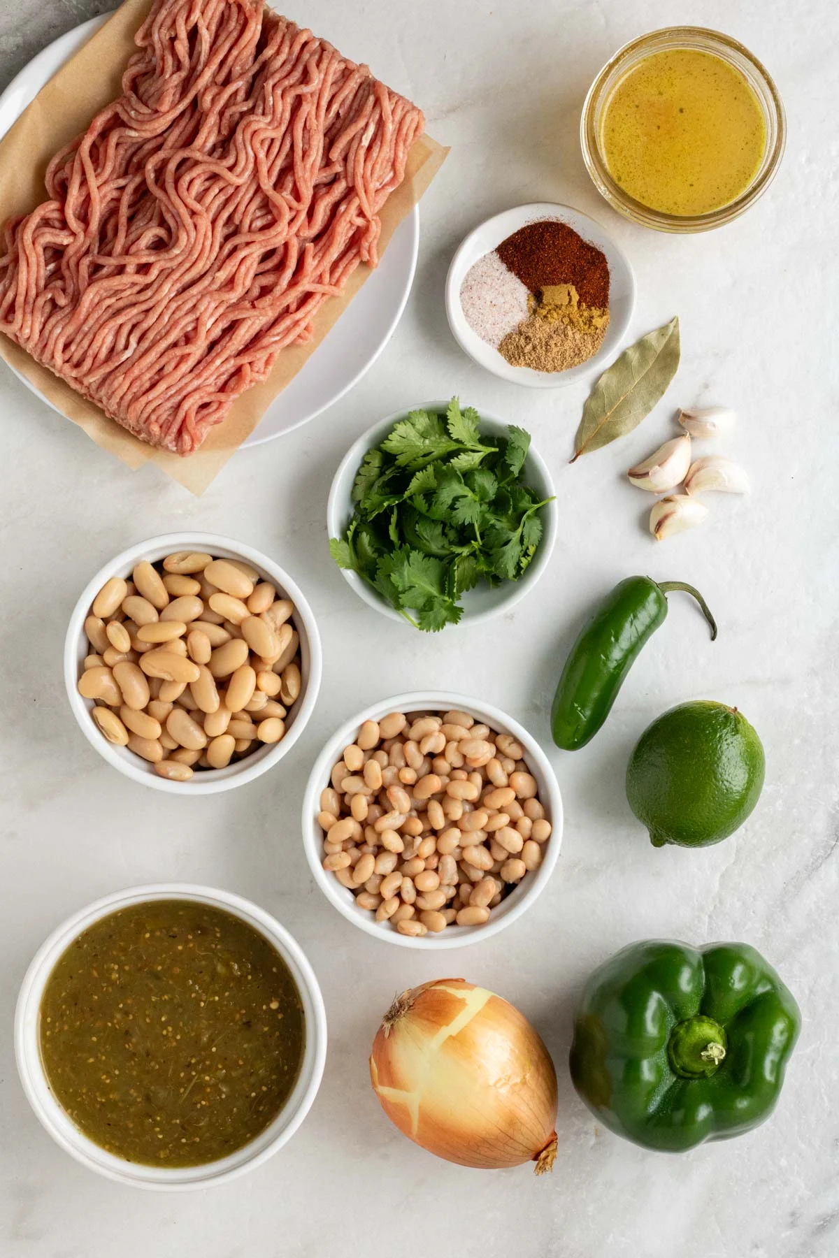 Plate of lean ground turkey, jar of chicken broth, bowls of spices, fresh cilantro, white beans, and crushed tomatillos with an onion, green bell pepper, jalapeño, lime, bay leaf, and garlic cloves on a white background.