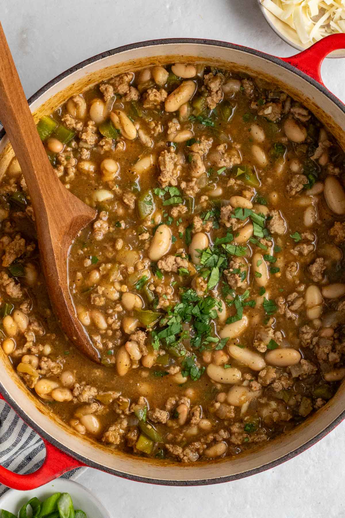 Large red pot of cooked turkey white bean chili with a wooden serving spoon and freshly chopped cilantro on top on a white background.