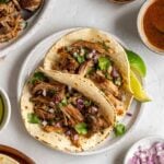Two slow-cooker pulled pork tacos on a speckled white plate with fresh cilantro and diced red onion on top and lime wedges on the side on a white background.