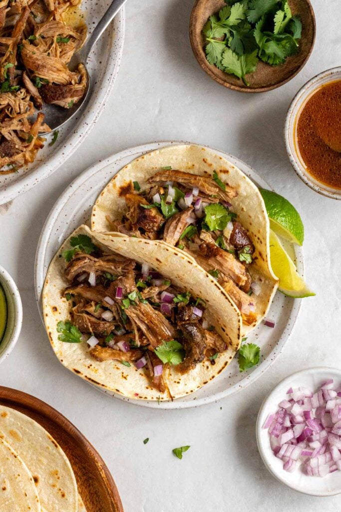 Two slow-cooker pulled pork tacos on a speckled white plate with fresh cilantro and diced red onion on top and lime wedges on the side on a white background.