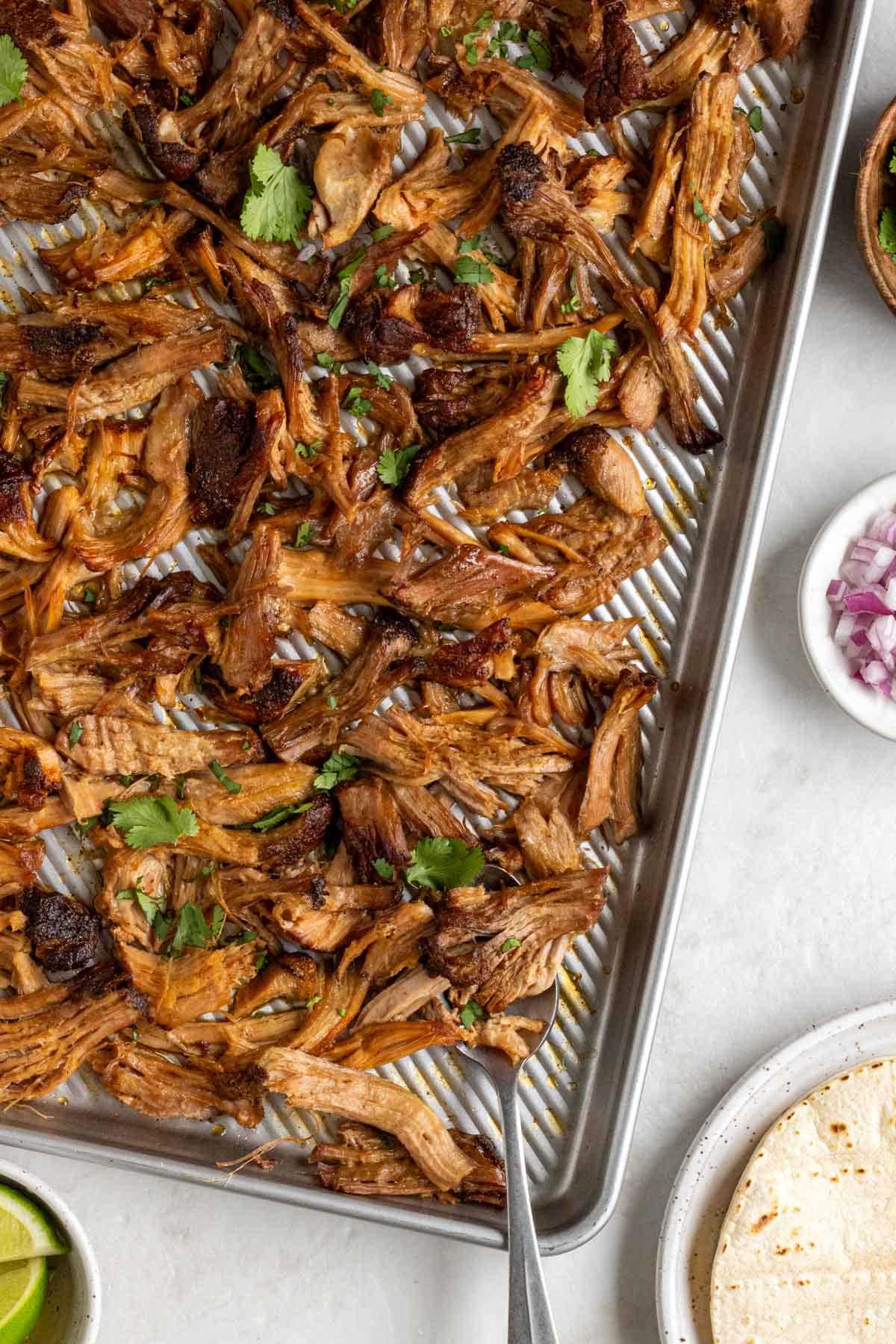 Baking sheet of crispy pulled pork with fresh cilantro on top with a silver serving spoon and a bowl of diced red onion, a bowl of lime wedges, and a plate of corn tortillas on the side on a white background.
