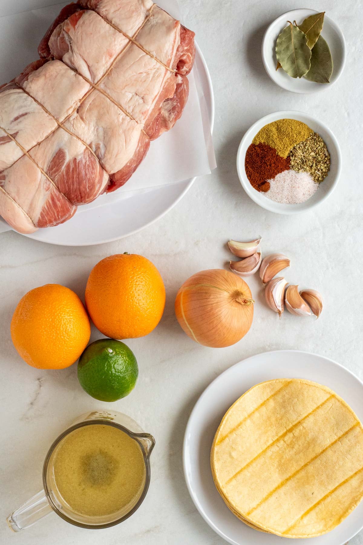 Large white plate with a raw pork shoulder on top with a bowl of bay leaves, a bowl of spices, two oranges, lime, onion, garlic cloves, glass jug of vegetable broth, and a plate of tortillas on a white background.