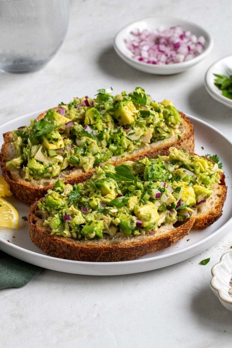 Plate of avocado tuna salad on toast with sliced lemon, diced red onion, parsley, and salt and pepper on the side.