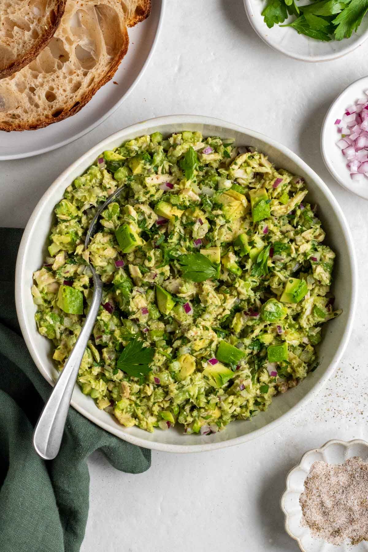 A bowl of tuna avocado salad with a silver serving spoon on a green tea towel with sourdough, parsley, onion, and salt on the side on a white background.