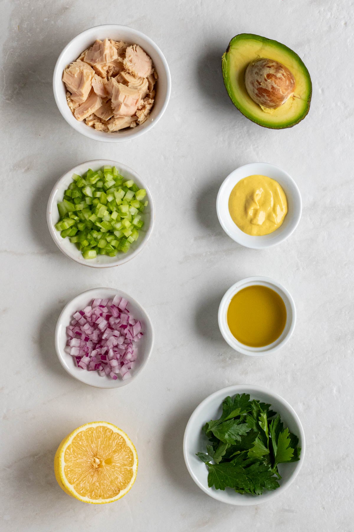 Bowl of tuna, half an avocado, bowl of diced celery, bowl of Dijon mustard, bowl of diced red onion, bowl of olive oil, half a lemon, and a bolw of fresh parsley on a white background.