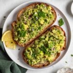 Two sliced of sourdough toast with avocado tuna salad on top on a plate with a dark green tea towel on a white background.