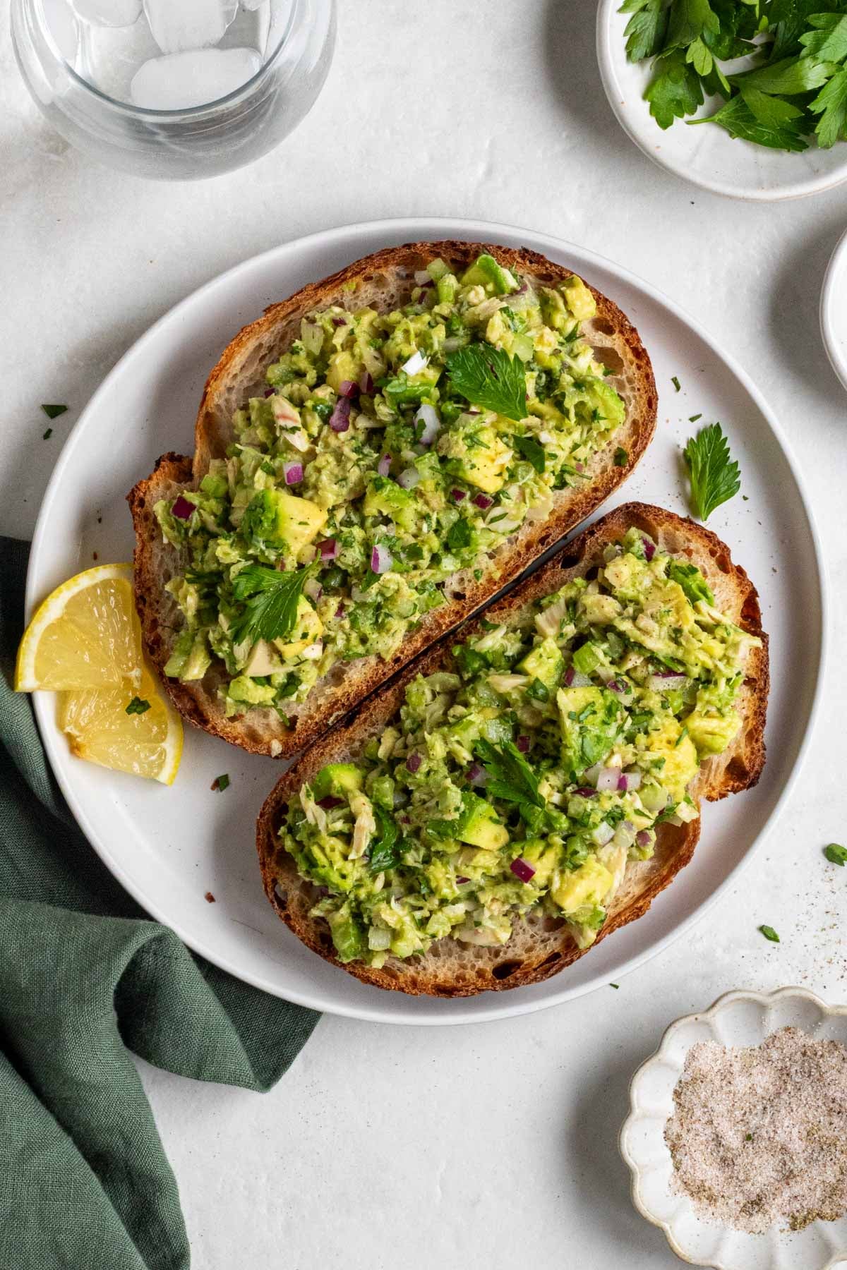 Two sliced of sourdough toast with avocado tuna salad on top on a plate with a dark green tea towel on a white background.