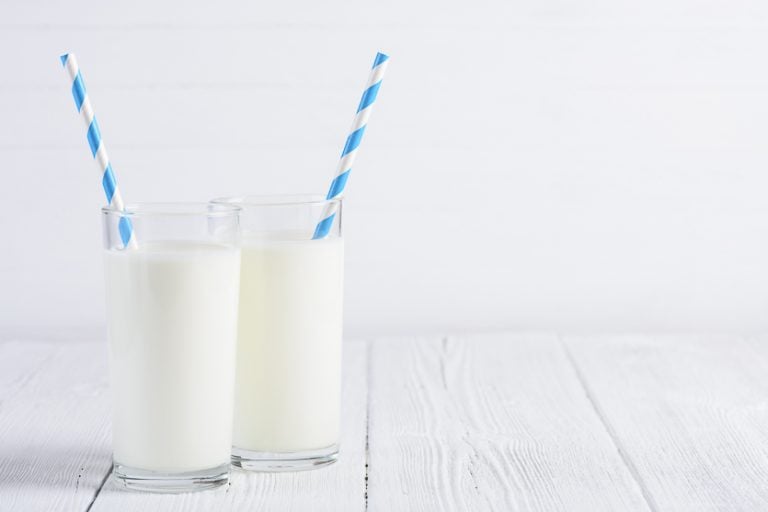 A glass of almond milk and cow's milk with striped blue and white straws on a white wood table on a white background.