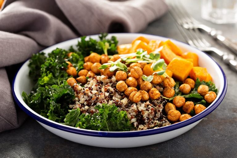 White bowl with kale, tri-color quinoa, roasted chickpeas, and roasted squash on a dark background with a grey tea towel, utensils, and a glass of water.