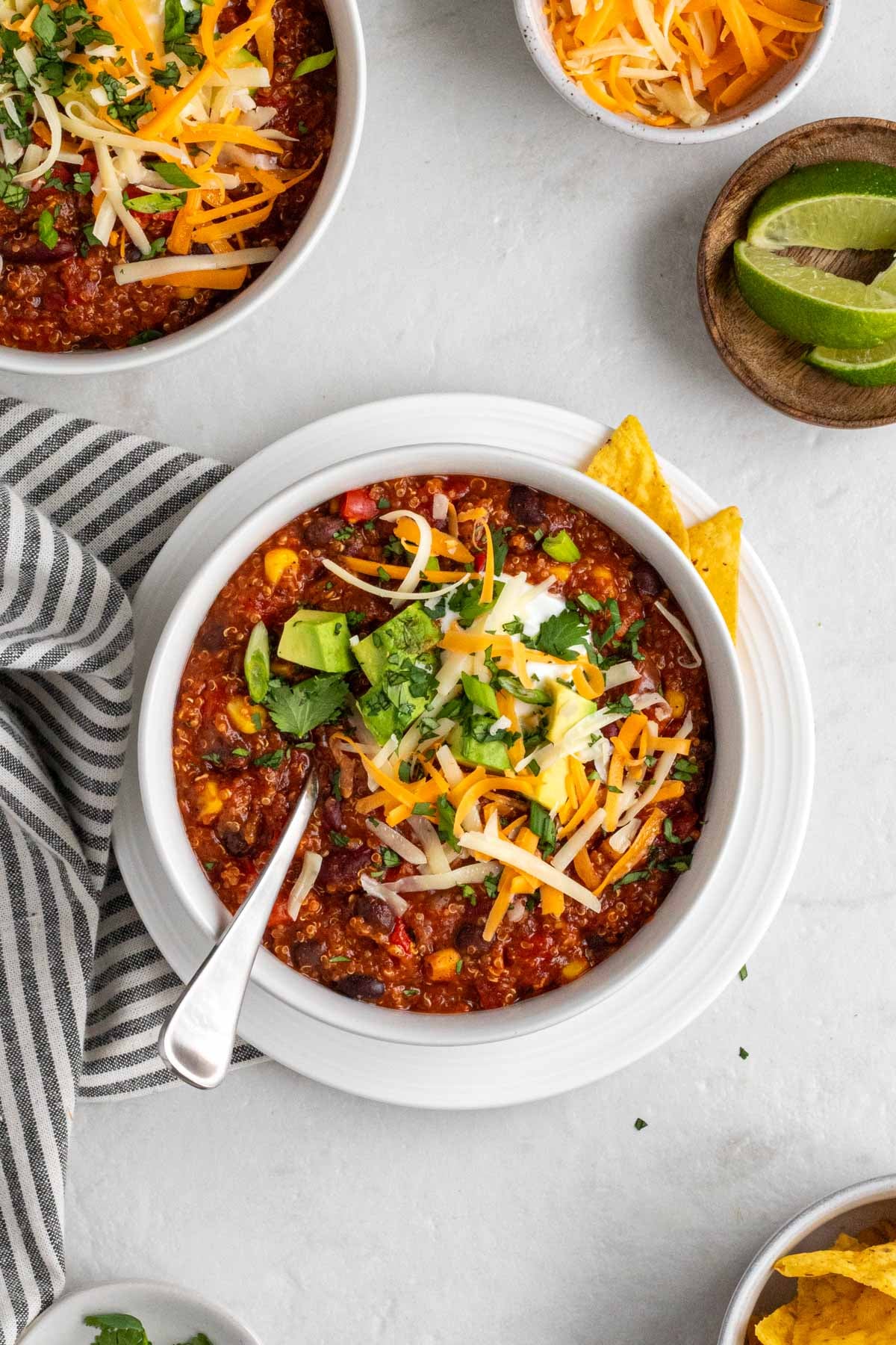 Two bowls of slow cooker vegetarian chili with sour cream, grated cheddar cheese, cubed avocado, sliced green onions, and chopped cilantro on top with tortilla chips, lime wedges, and a tea towel on the side on a white background.