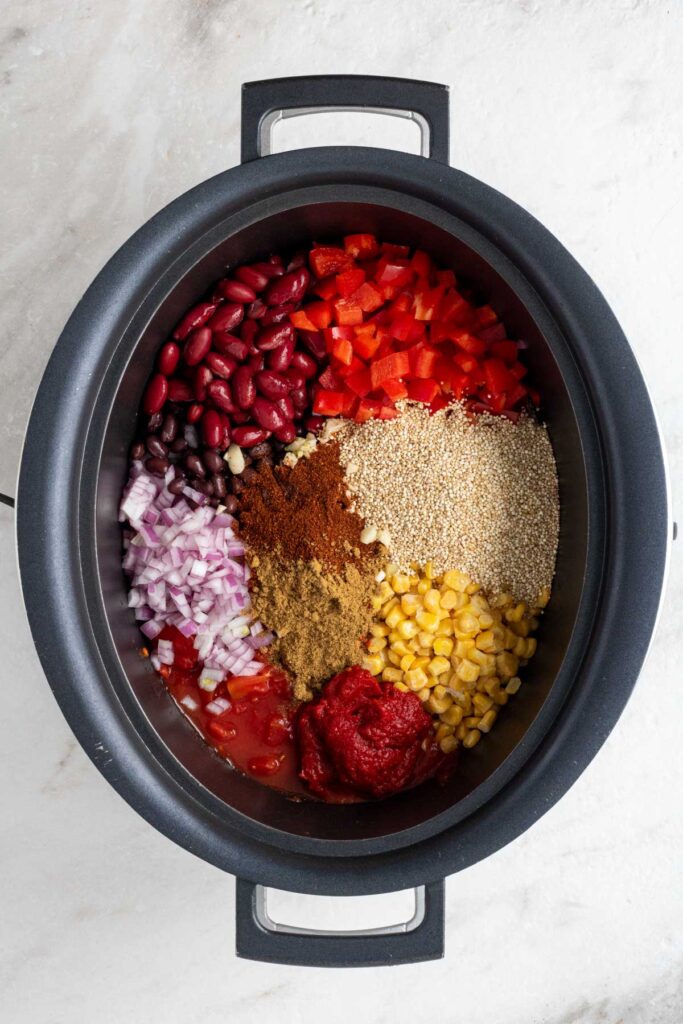 Slow cooker filled with diced red bell pepper, quinoa, frozen corn, tomato paste, diced tomatoes, diced red onion, black beans, red kidney beans, minced garlic, and spices on a white background.