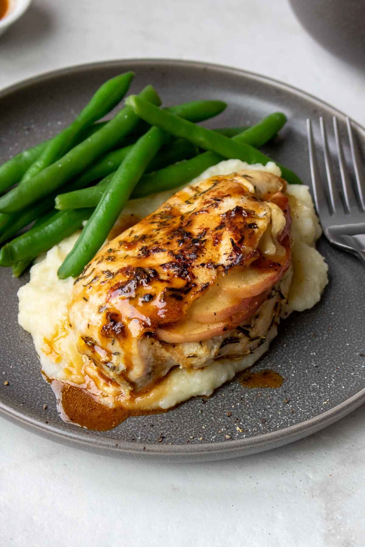 Apple brie stuffed chicken breast covered in sauce with mashed potatoes and green beans on a grey plate with a fork.