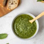 Small bowl of fresh basil pesto with a wooden spoon with a side of crusty bread and basil leaves.