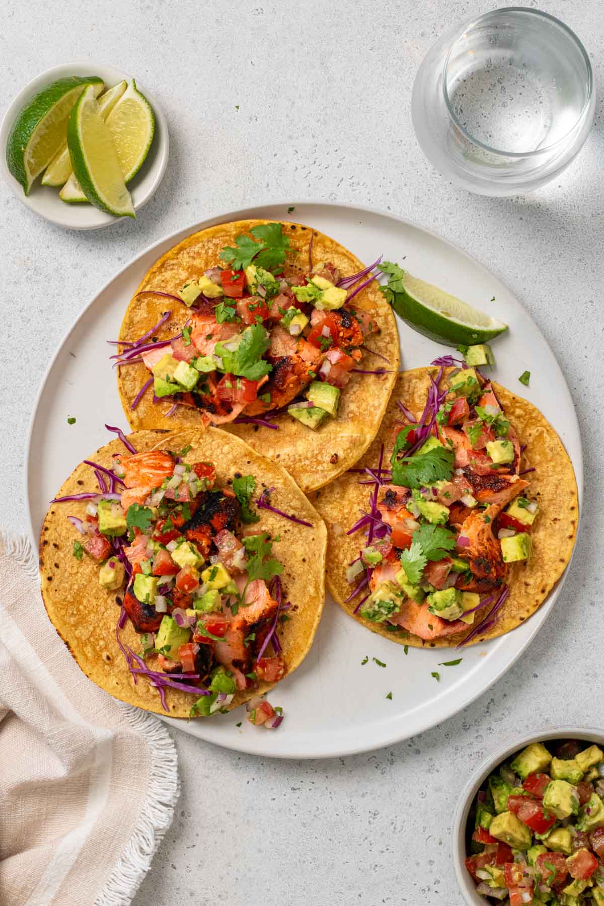 Plate with three grilled salmon tacos with a side of avocado salsa, cilantro, and lime wedges.
