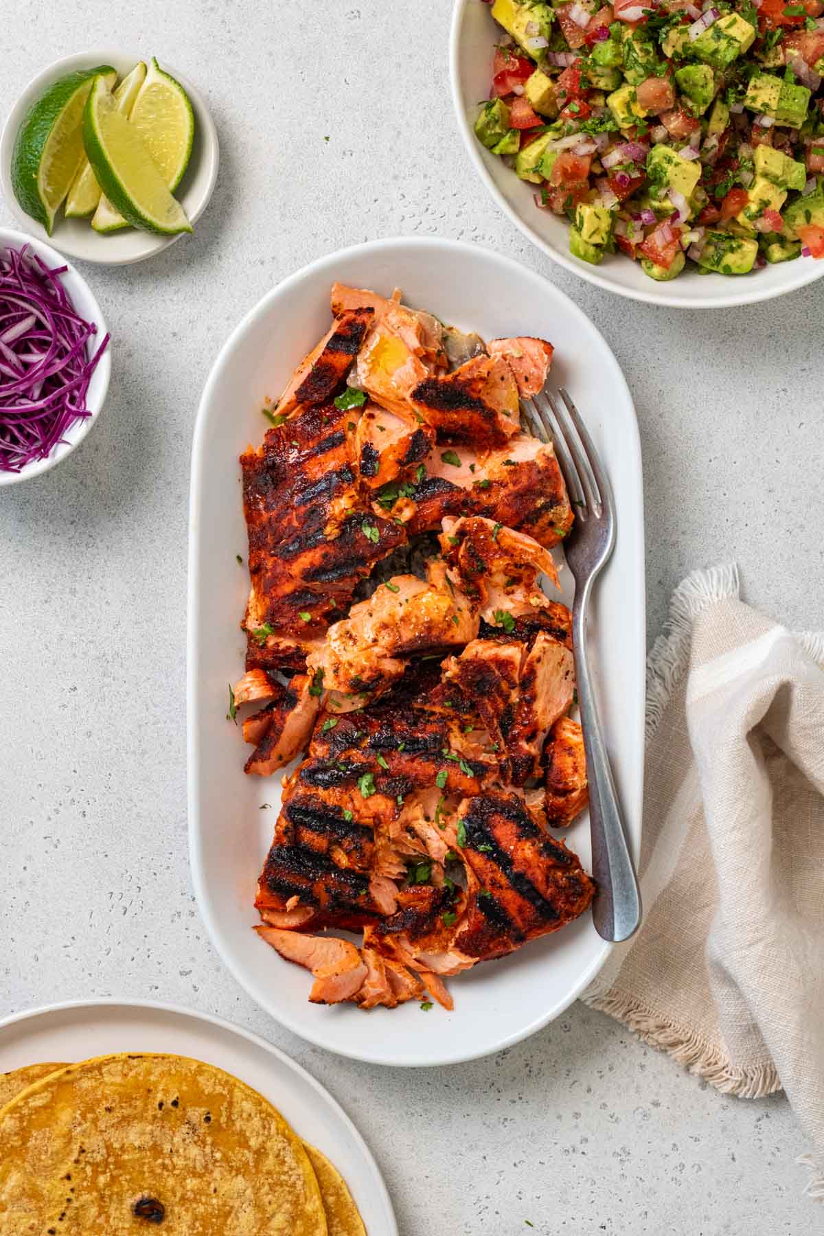 Serving dish with grilled salmon with a bowl of avocado salsa, a plate of corn tortillas, a bowl of shredded cabbage, and lime wedges.