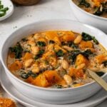 White bowl of butternut squash, kale, and white bean soup with a spoon with a side of crusty bread, parmesan cheese, and parsley.