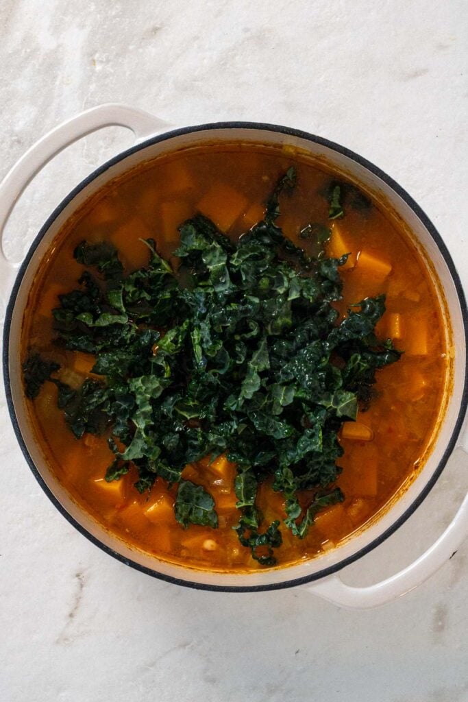 Large pot with sautéed onions, spices, butternut squash, white beans, parmesan cheese, broth, and chopped kale.