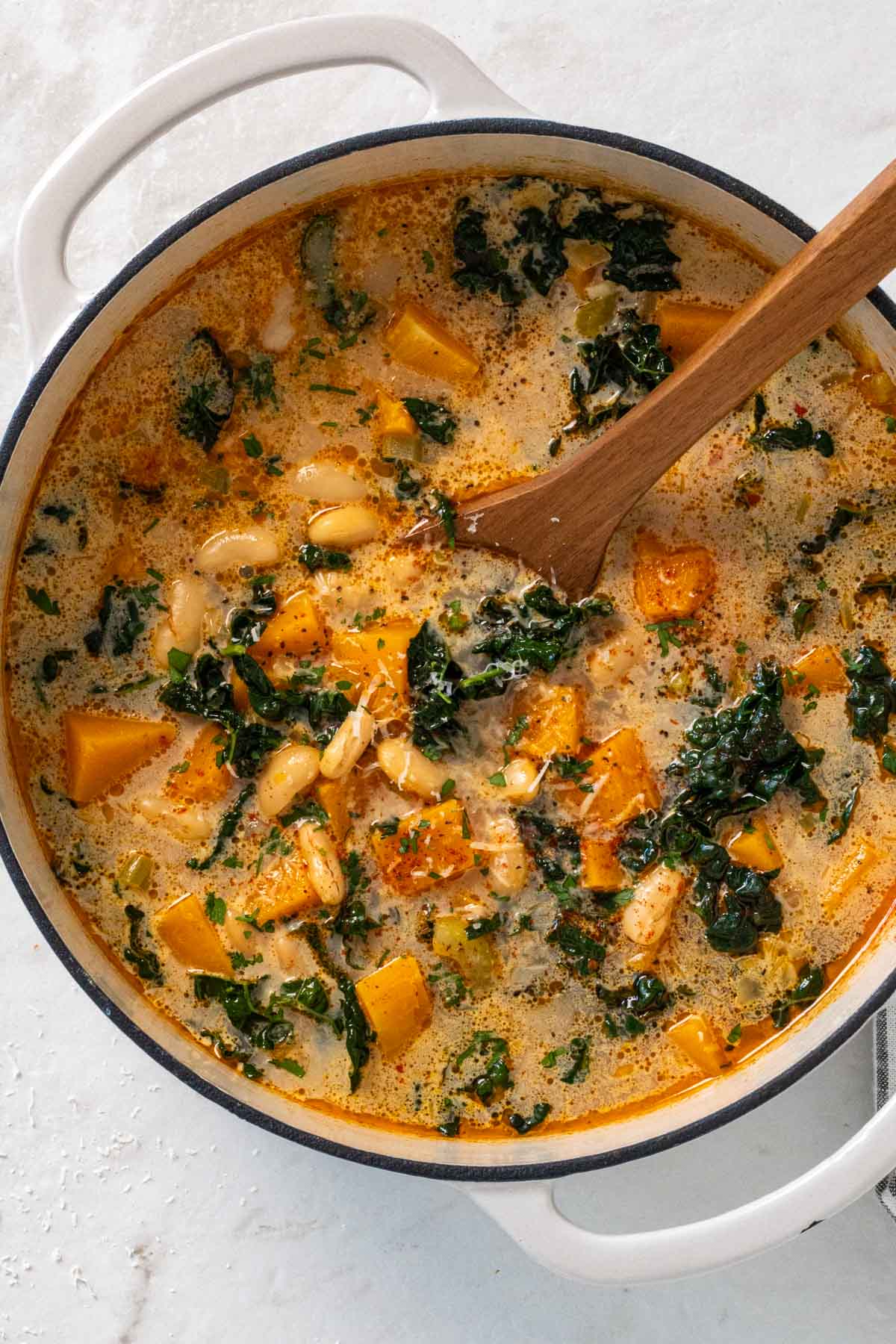 Large pot of cooked butternut squash, kale, and white bean soup with chopped parsley and parmesan cheese on top.