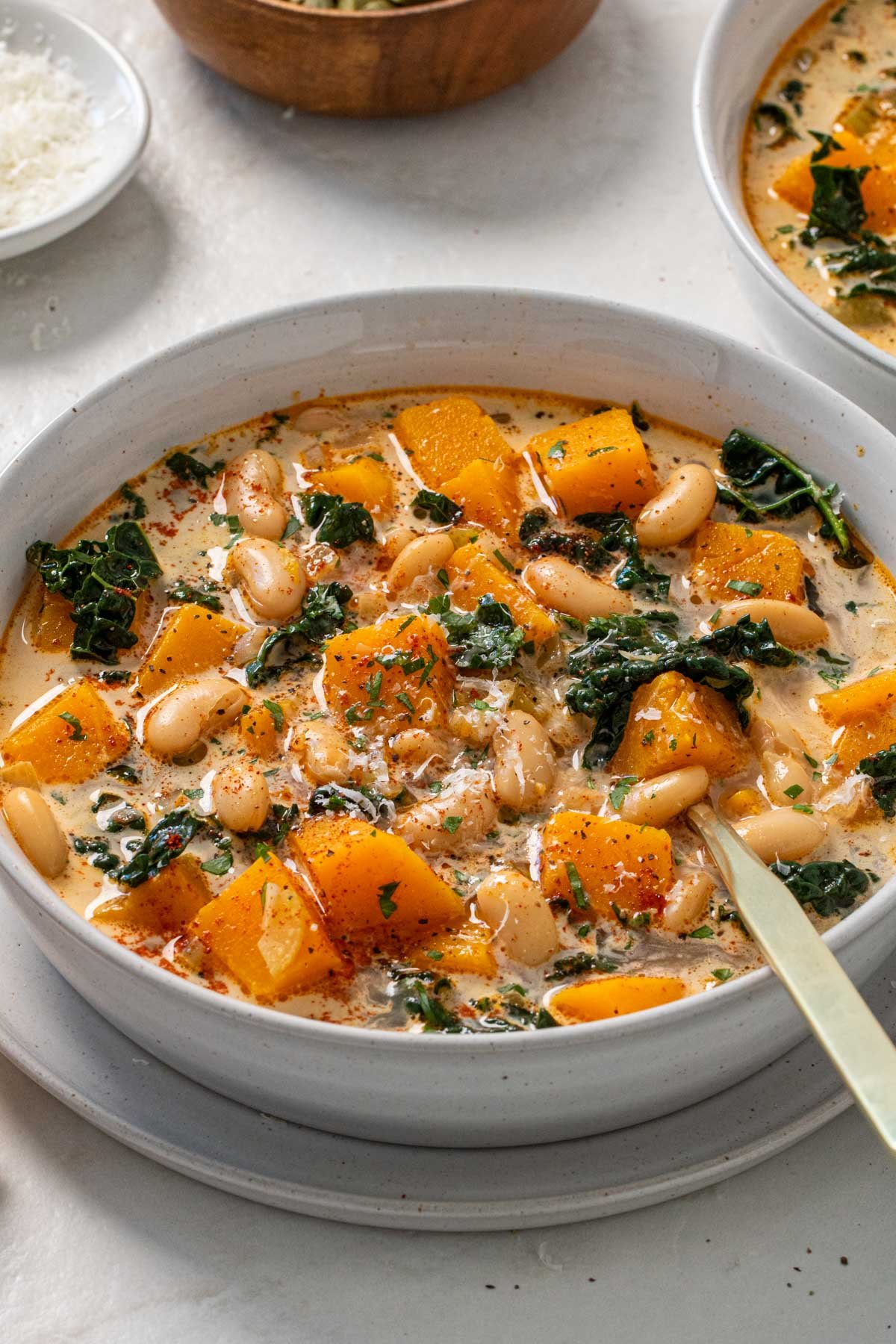 Close up of a bowl of kale, butternut squash, and white bean soup with a spoon and parmesan cheese and toasted pumpkin seeds on top.