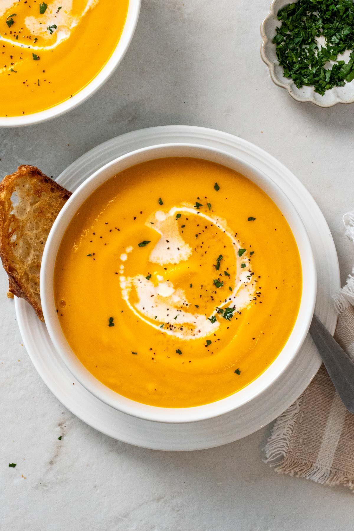 Two bowls of carrot parsnip soup with crusty bread on a white plate with a spoon and tea towel with a drizzle of cream, parsley, and fresh black pepper on top.