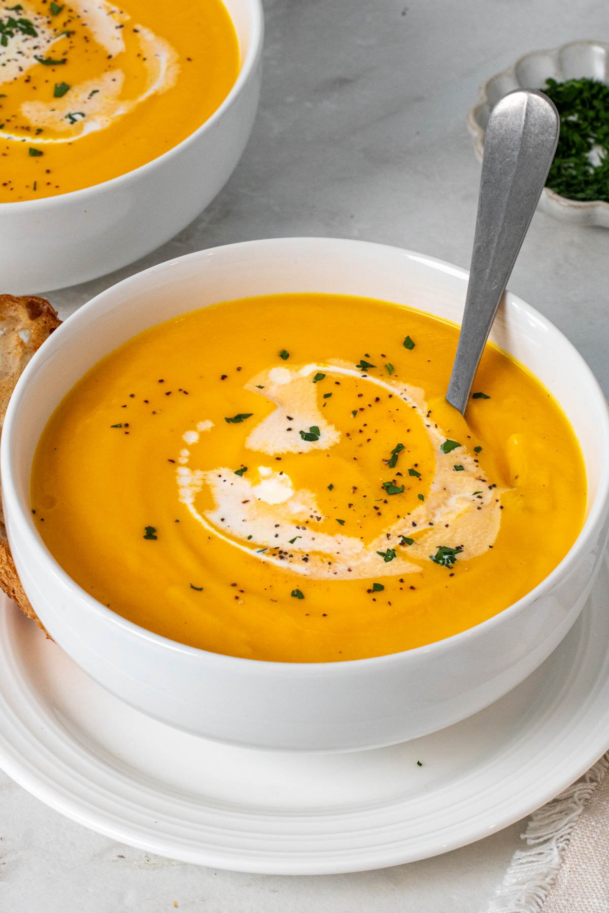 White bowl with carrot parsnip soup with chopped parsley on top and a spoon with a side of crusty bread.