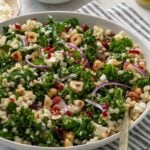 Bowl of couscous kale salad with feta cheese, cranberries, and hazelnuts on a stripped green tea towel with a gold spoon.