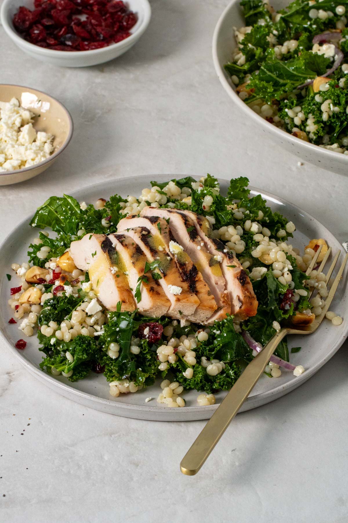 Plate of kale couscous salad with grilled chicken on top with a fork and a side of crumbled feta cheese and dried cranberries.