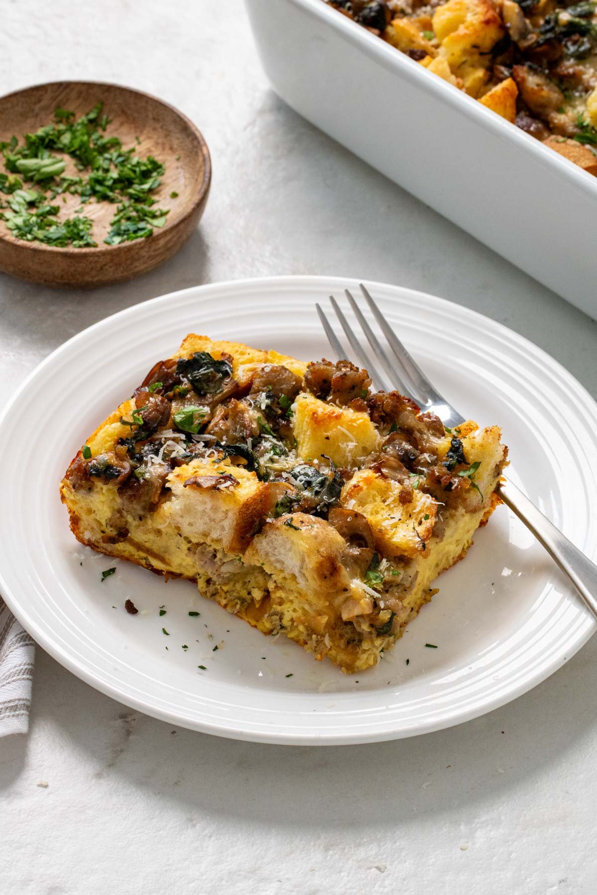 Slice of bread, egg, and sausage breakfast casserole on a while plate with a fork and a side of fresh parsley.