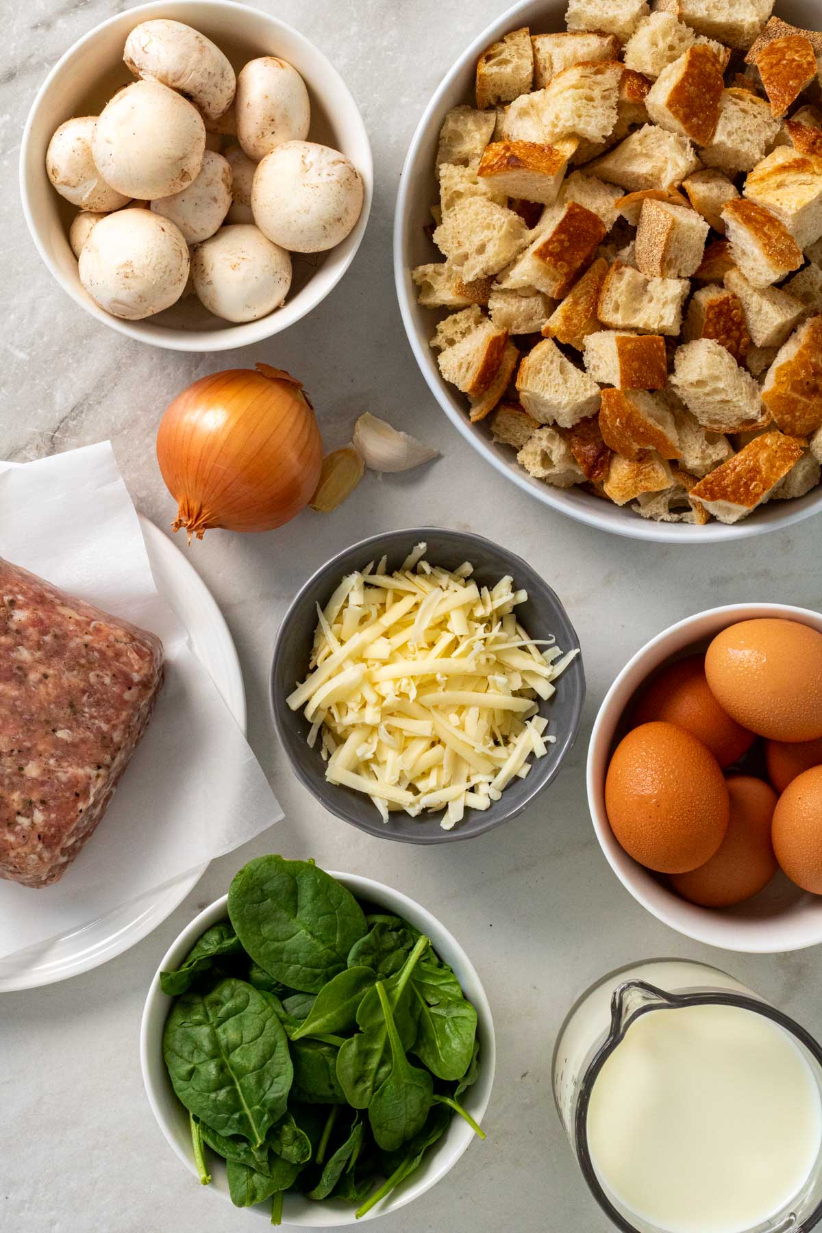 Ingredients for bread, egg, and sausage breakfast casserole: white bread, pork sausage, eggs, milk, cheddar cheese, onion, garlic, mushrooms, and spinach.