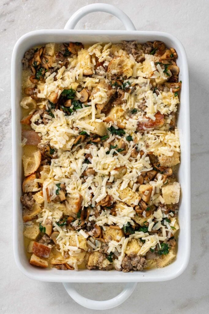 Baking dish with bread cubes, pork sausage, vegetables, egg mixture, and shredded cheese on top.