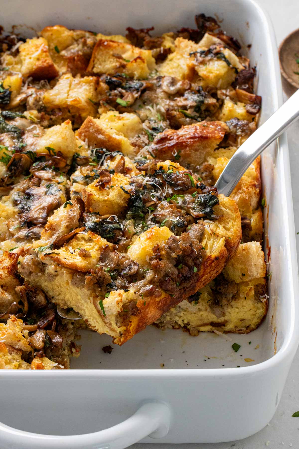 Slice of egg, sausage, and bread breakfast casserole recipe on a spatula in a white baking dish.