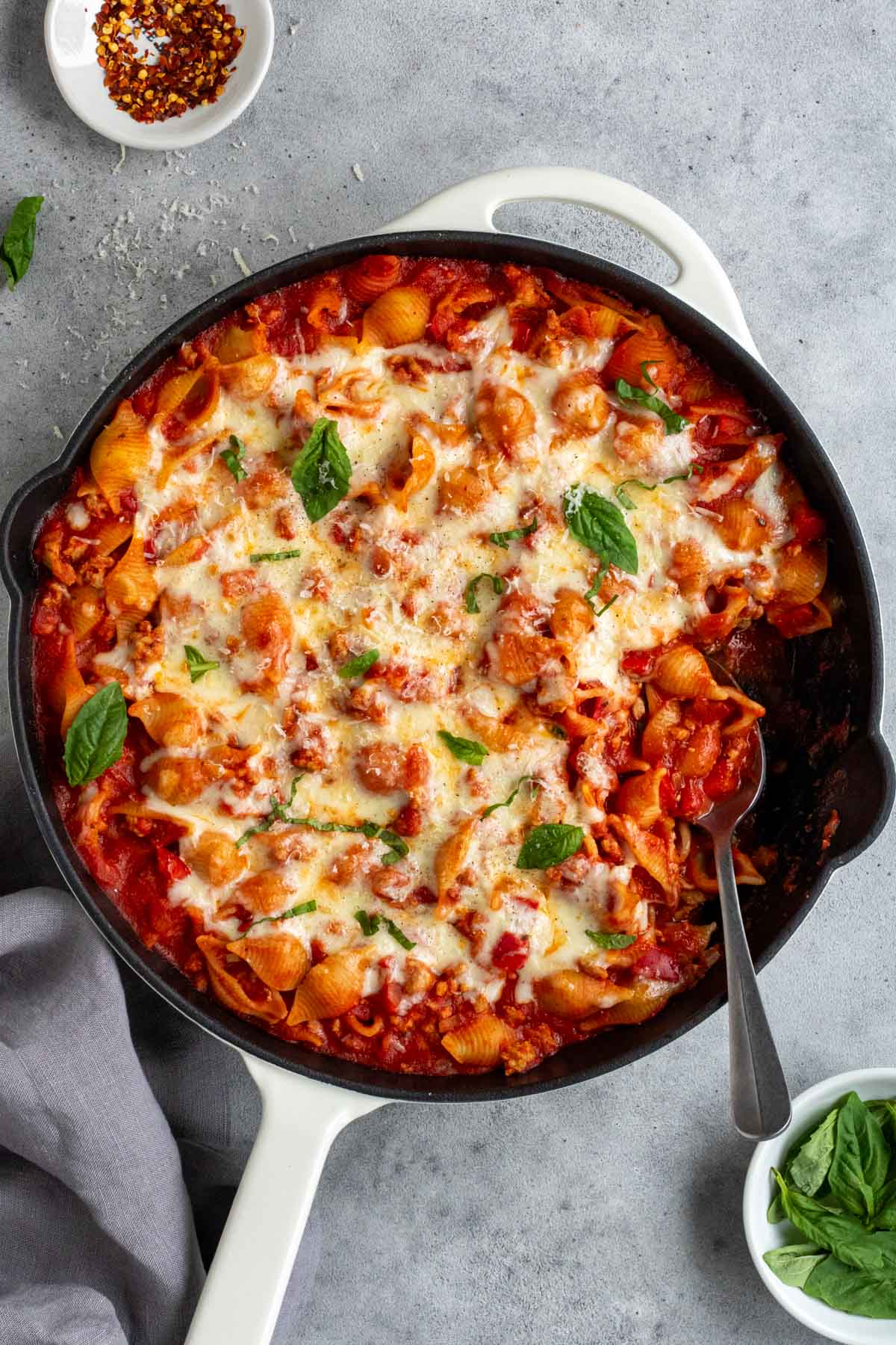 Ground turkey pasta bake in a cast-iron skillet with fresh basil on top, a serving spoon, and a side of crushed red pepper flakes.